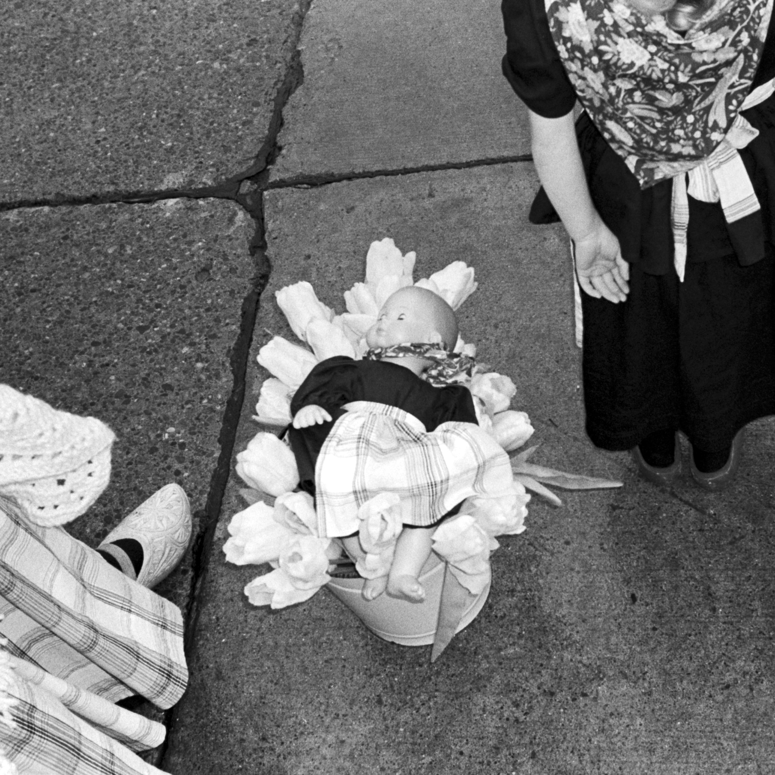 A doll dressed in traditional clothing lying among flower petals on the sidewalk, with a child standing nearby and only part of another person visible. Pella, Iowa Tulip Time Festival, Dutch Heritage