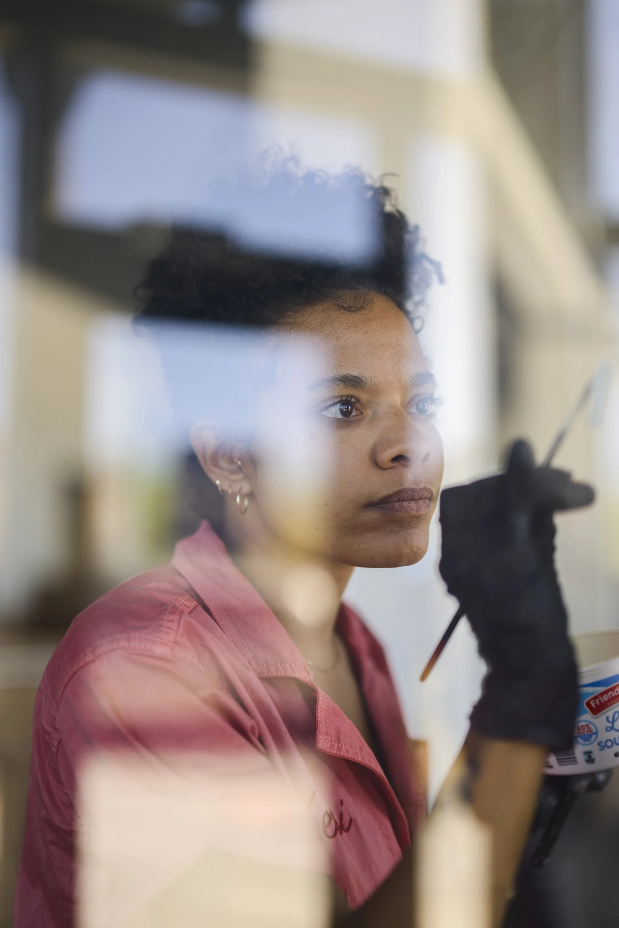 A young artist painter woman with natural curly hair and earrings, wearing a pink shirt, looking thoughtfully through a glass window while holding a paintbrush and paint container