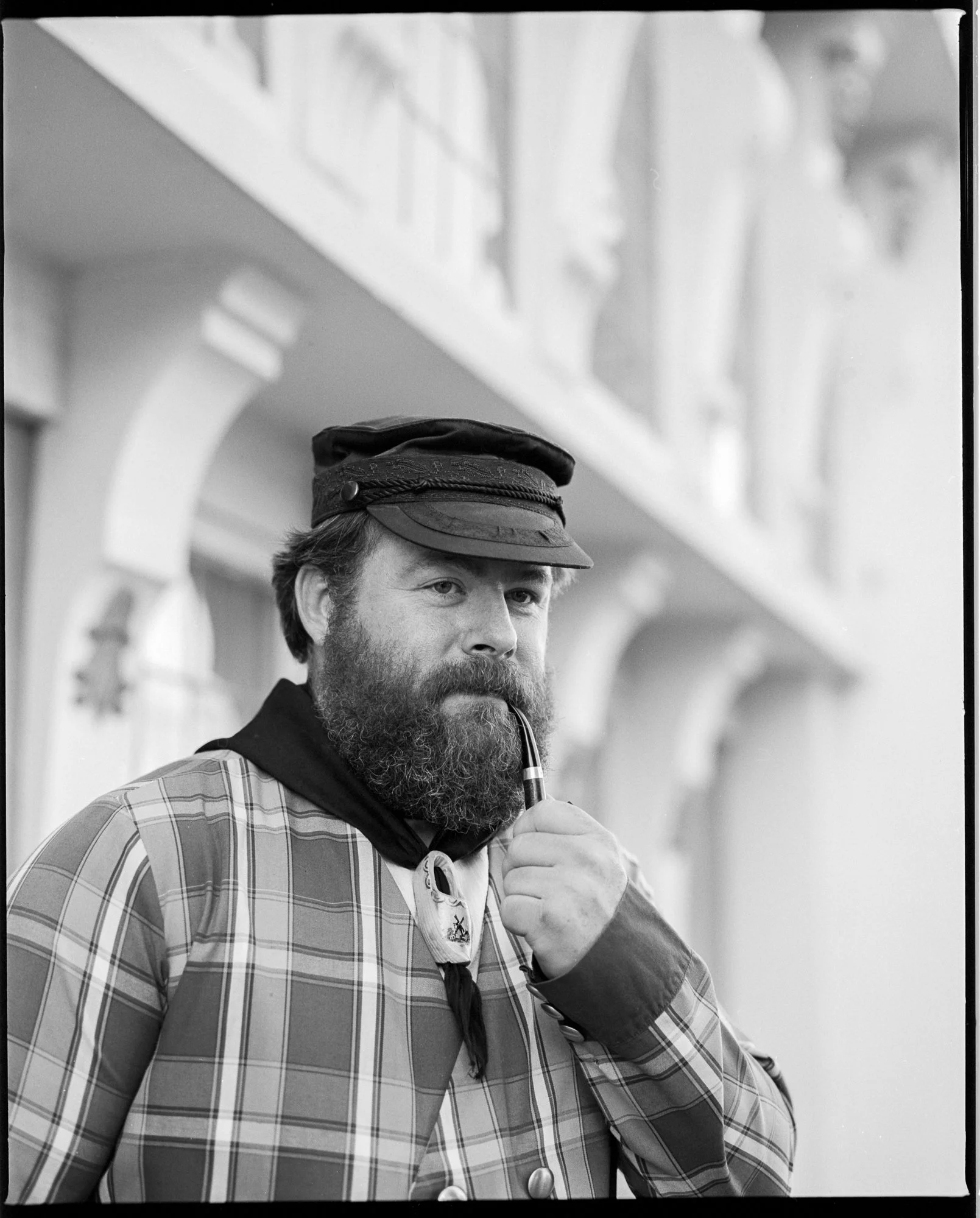 A man with a beard and mustache wearing a cap and plaid jacket, holding a pipe near his mouth, standing in front of a building with arches. Pella, Iowa Tulip Time Festival, Dutch Heritage