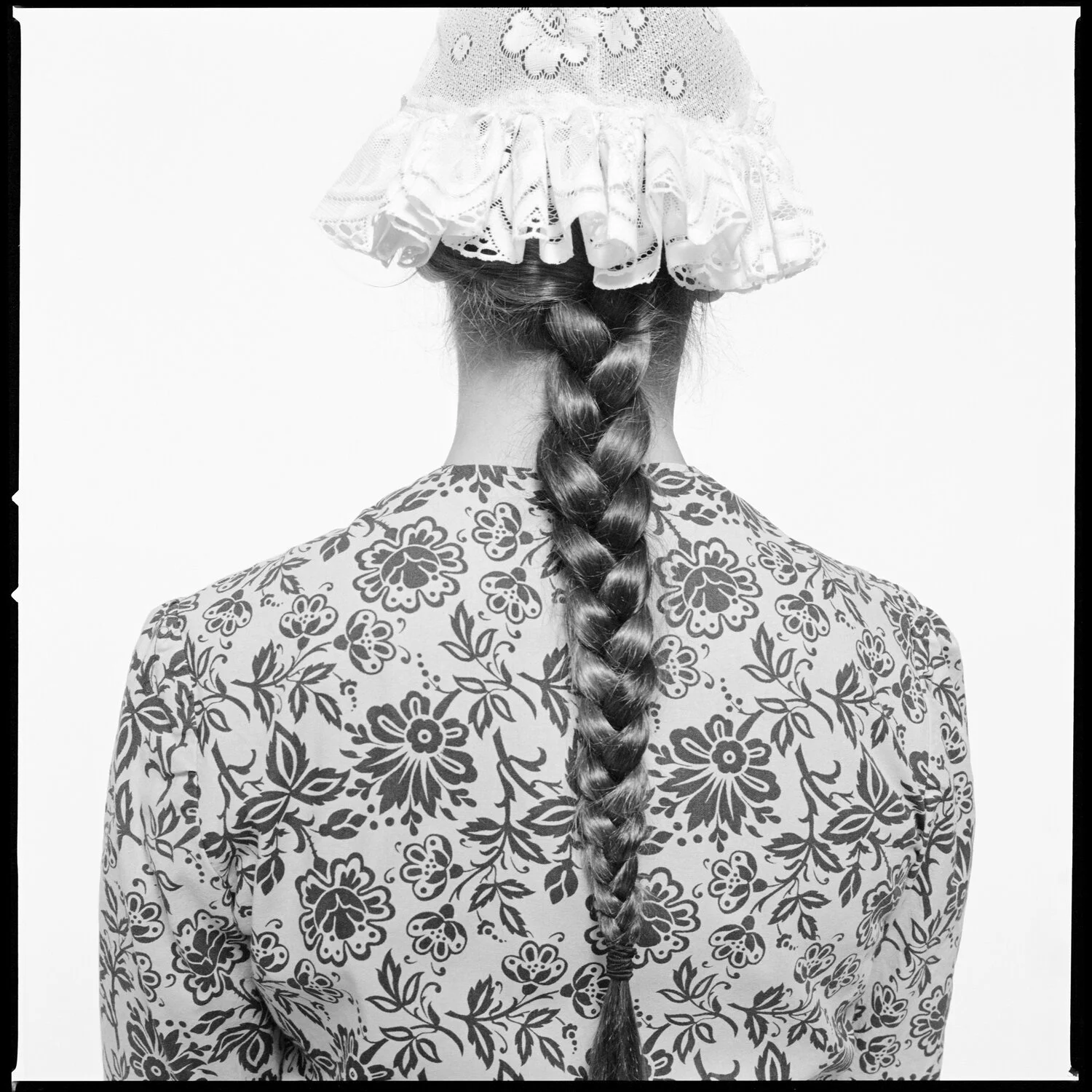 Back view of a woman with a detailed, floral patterned shirt, long braided hair, and a lacy, decorative hat with ruffles. Pella, Iowa Tulip Time Festival, Dutch Heritage