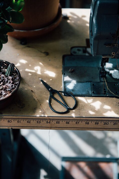 A pair of small black scissors resting on a wooden workbench with a ruler marked in inches, near a teal-colored sewing machine, with potted plants around.