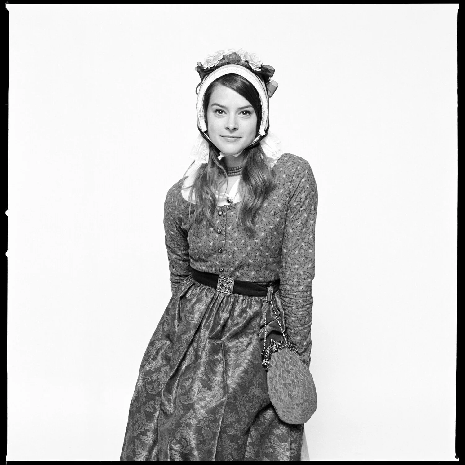A woman dressed in traditional European folk costume, wearing a bonnet and patterned skirt, standing against a plain background. Pella, Iowa Tulip Time Festival, Dutch Heritage