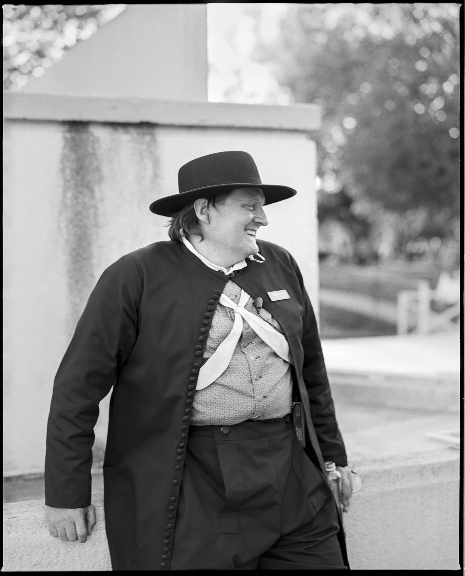 A man dressed in historical attire, smiling and walking outdoors near a building, wearing a wide-brimmed hat and a buttoned coat. Pella, Iowa Tulip Time Festival, Dutch Heritage