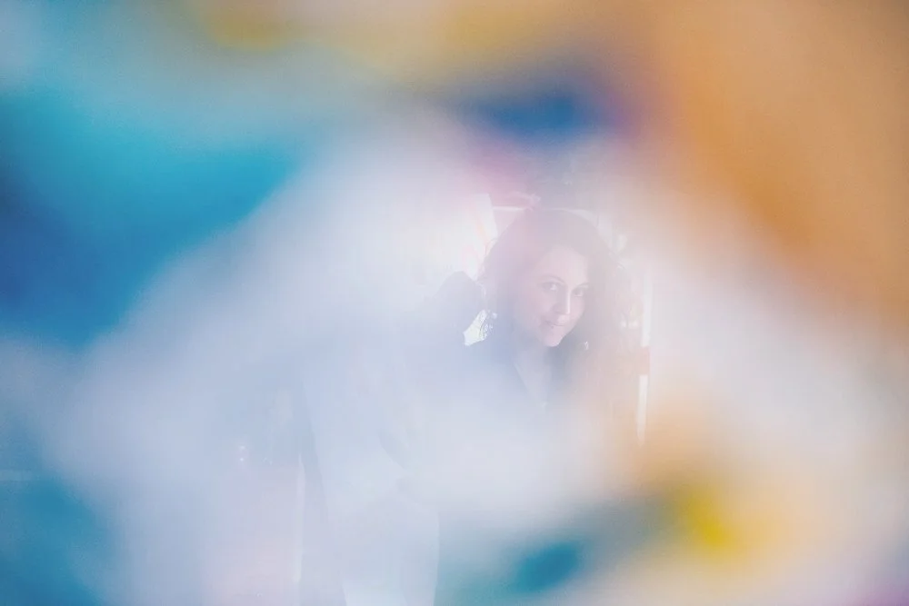 A woman painter artist with dark hair smiling, seen through colorful, blurry, and hazy foreground elements.