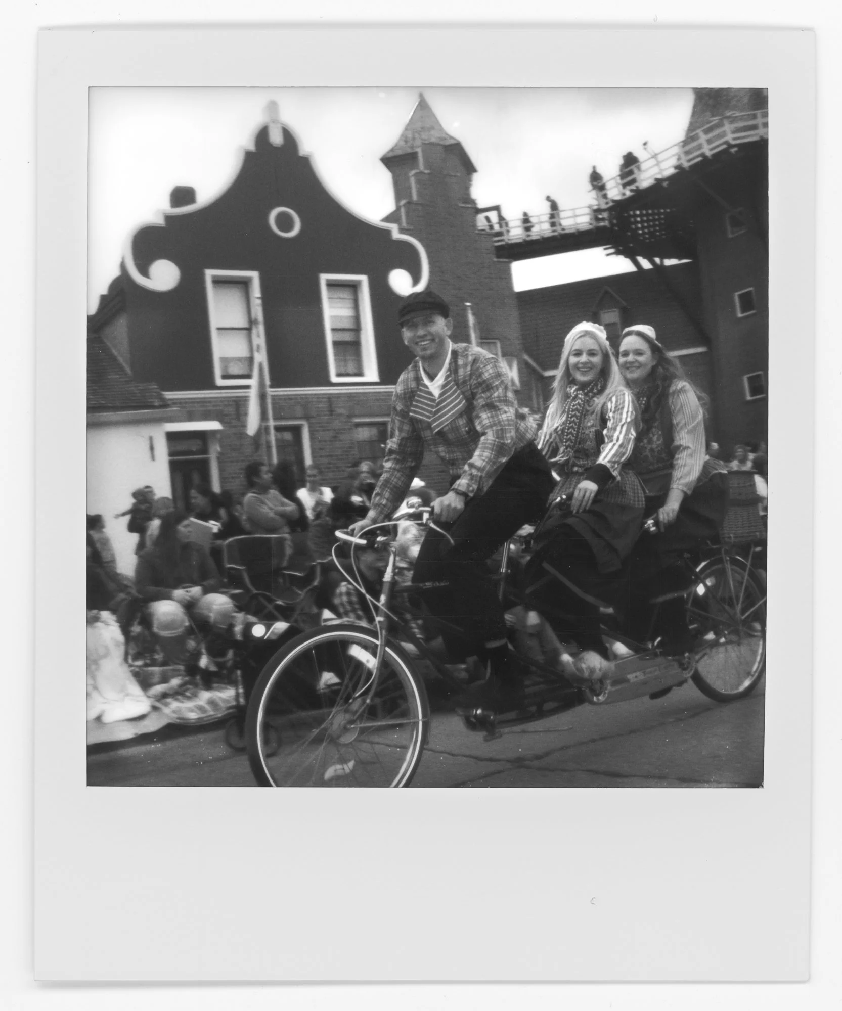 People riding a vintage bicycle with two women and a man in costumes in front of a festive building with decorative architecture. Pella, Iowa Tulip Time Festival, Dutch Heritage