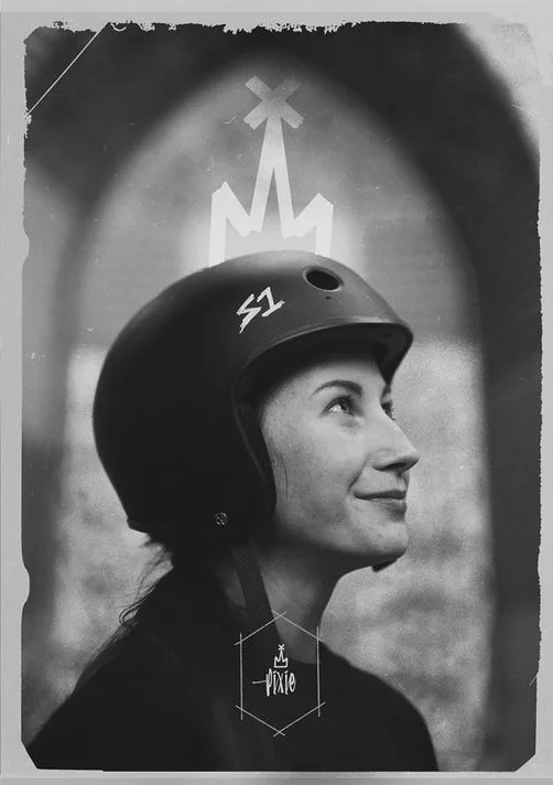 A woman skater in a helmet with the number 51 looks upward, with a background featuring a glowing arrow and a cross-like symbol. Sk8 Liborius