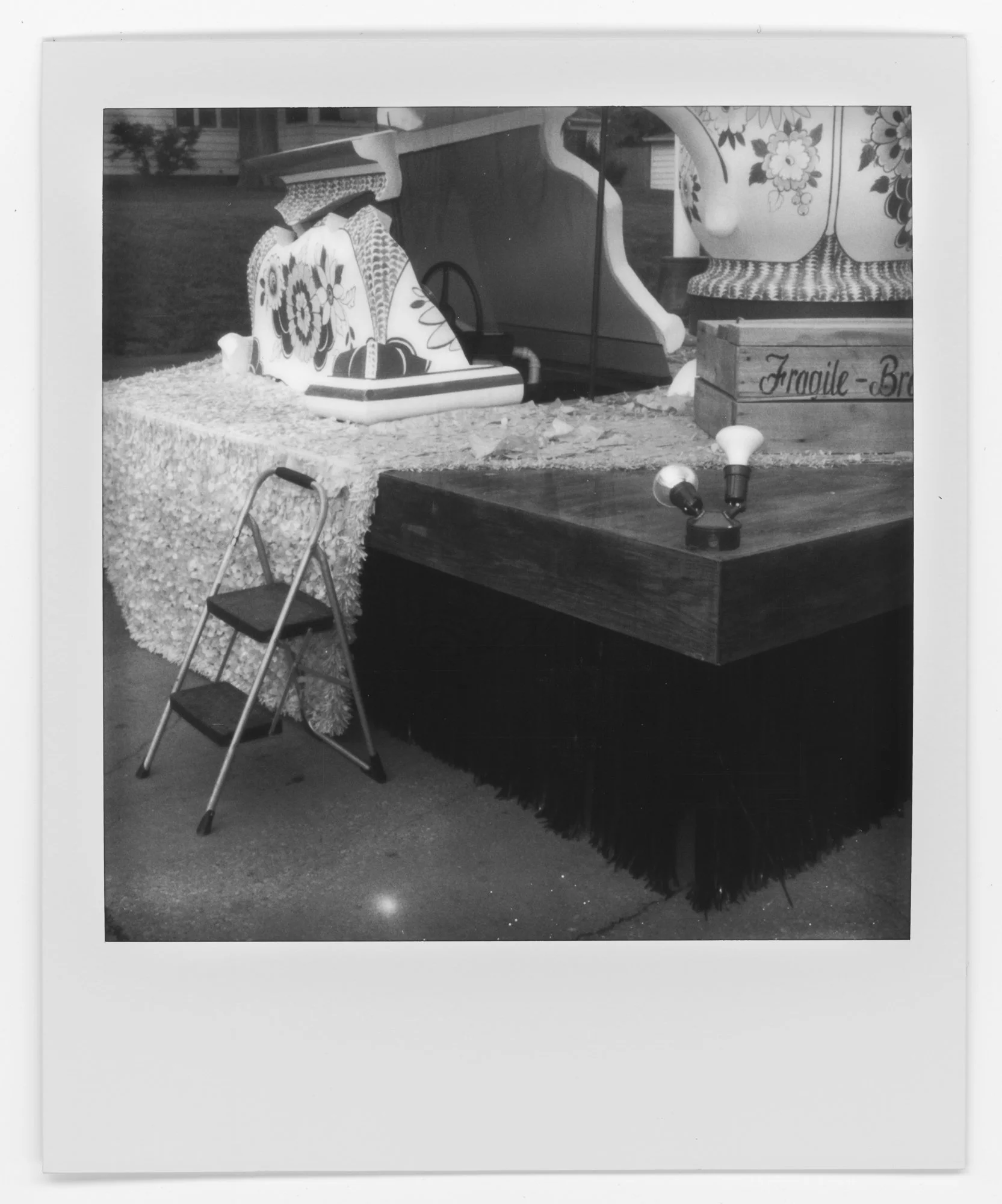 Black and white Polaroid photo of a toy setup with a small step ladder, a table with a lace cloth, and various decorative items including a teapot and a wooden box labeled 'Fragile-Brown'. Pella, Iowa Tulip Time Festival, Dutch Heritage