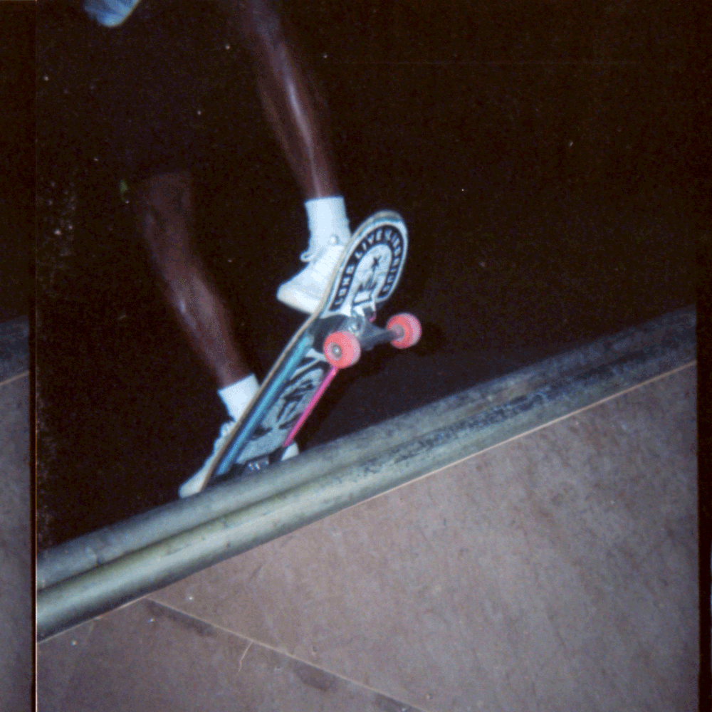 Close-up of a skateboard with pink wheels and a graphic design, being kicked up by a person wearing white sneakers and dark pants, riding over a black surface. Sk8 Liborius