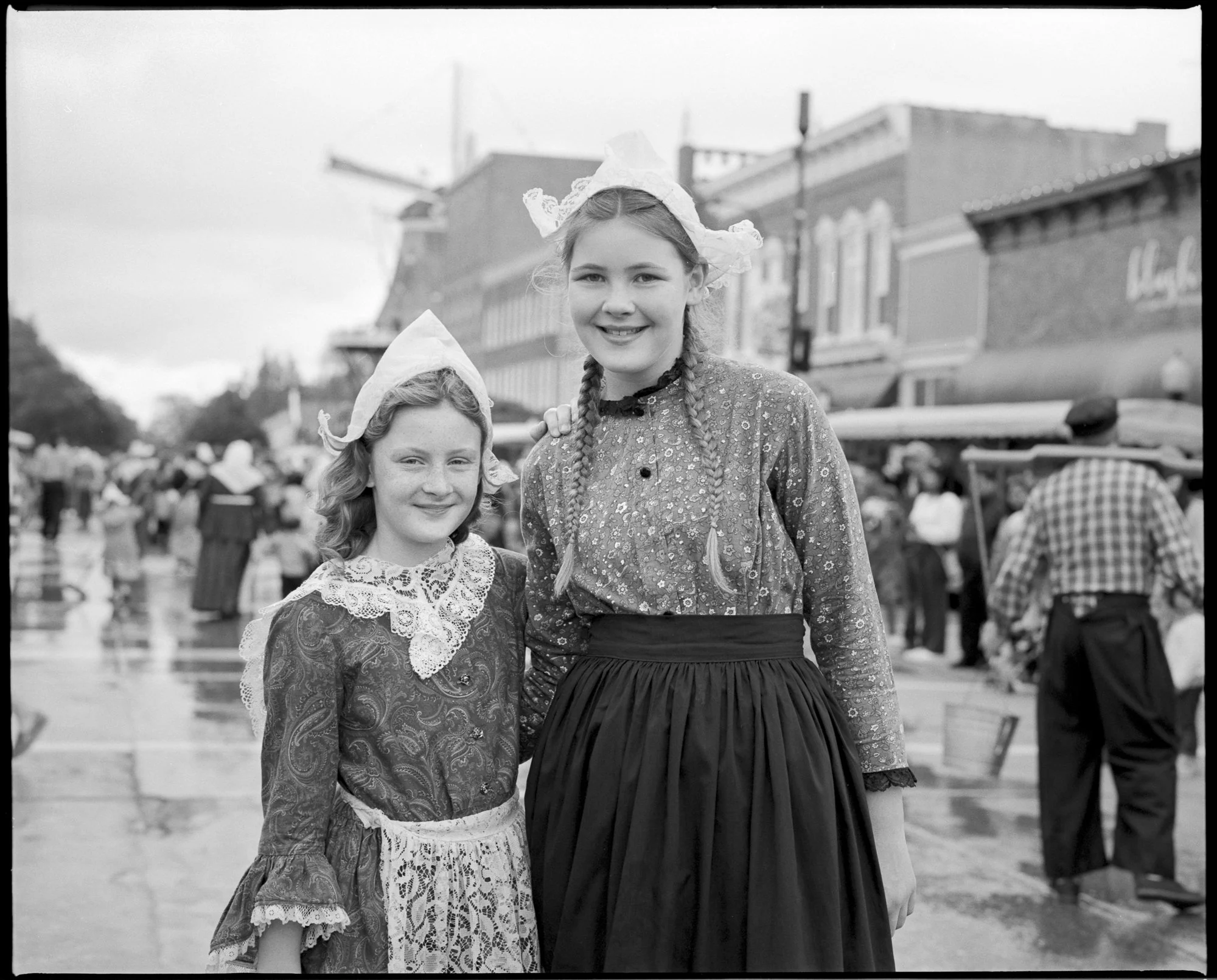 Two young women in historical attire, smiling, standing on a busy street during a festival or parade, with a crowd of people and historic buildings in the background. Pella, Iowa Tulip Time Festival, Dutch Heritage