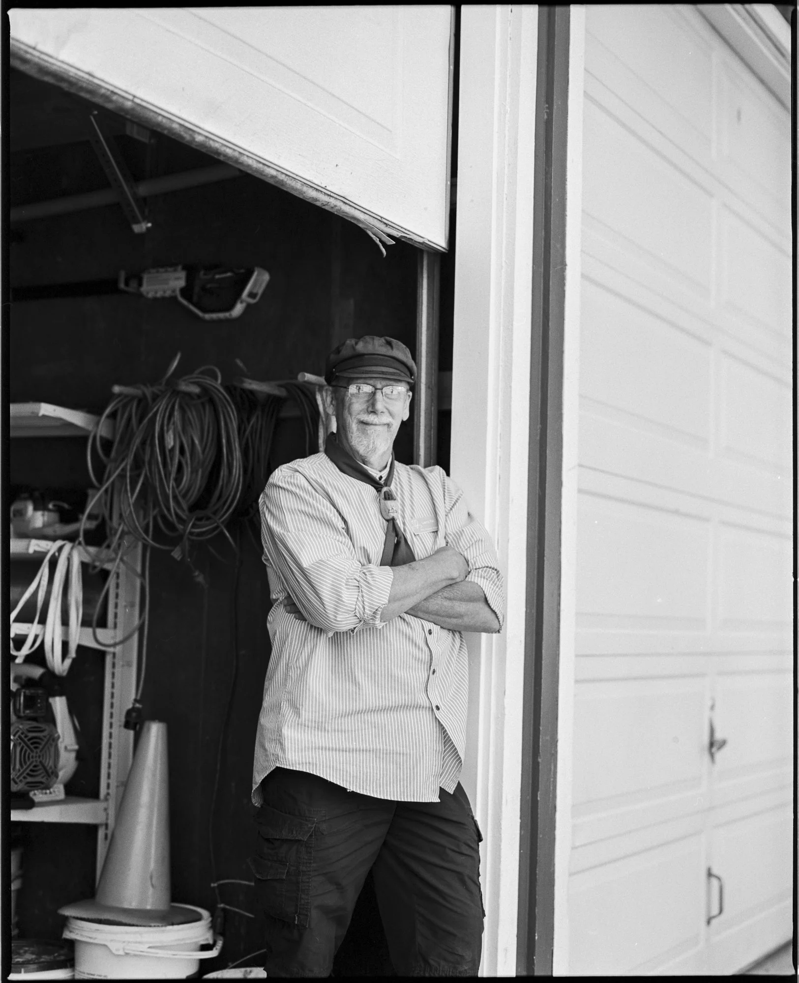 A man with glasses, a beard, and a cap standing with crossed arms inside a garage, with tools and coiled cords on shelves behind him. Pella, Iowa Tulip Time Festival, Dutch Heritage
