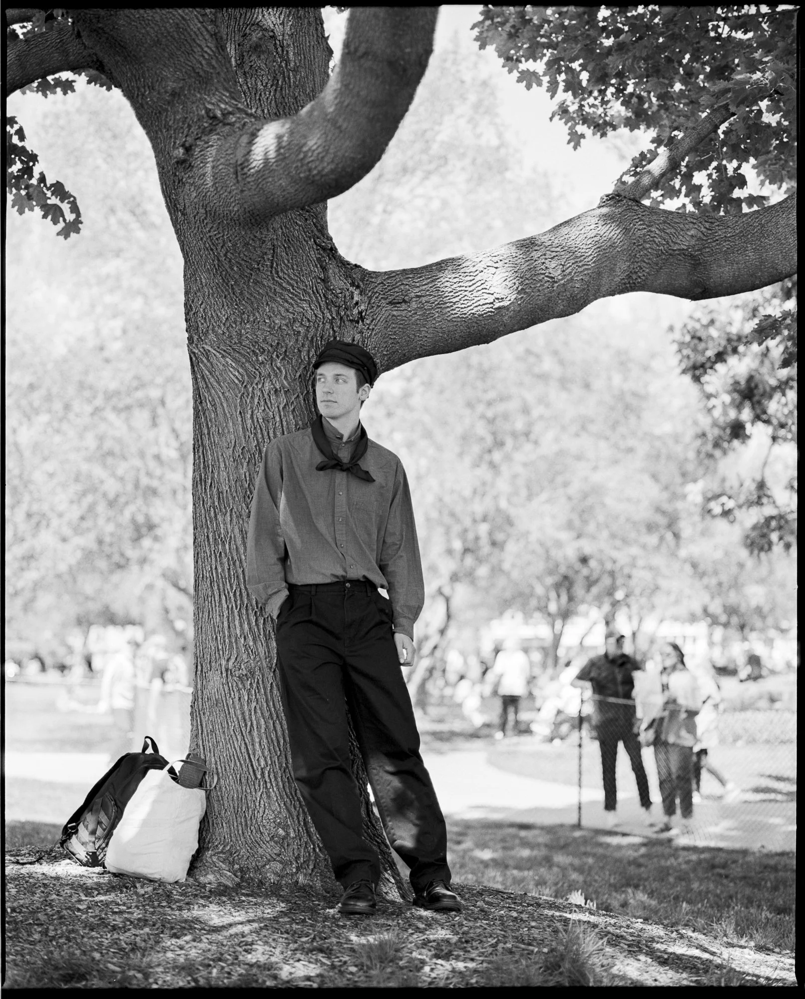 A young man in vintage clothing leaning against a large tree in a park, with a backpack on the ground nearby and people walking in the background. Pella, Iowa Tulip Time Festival, Dutch Heritage