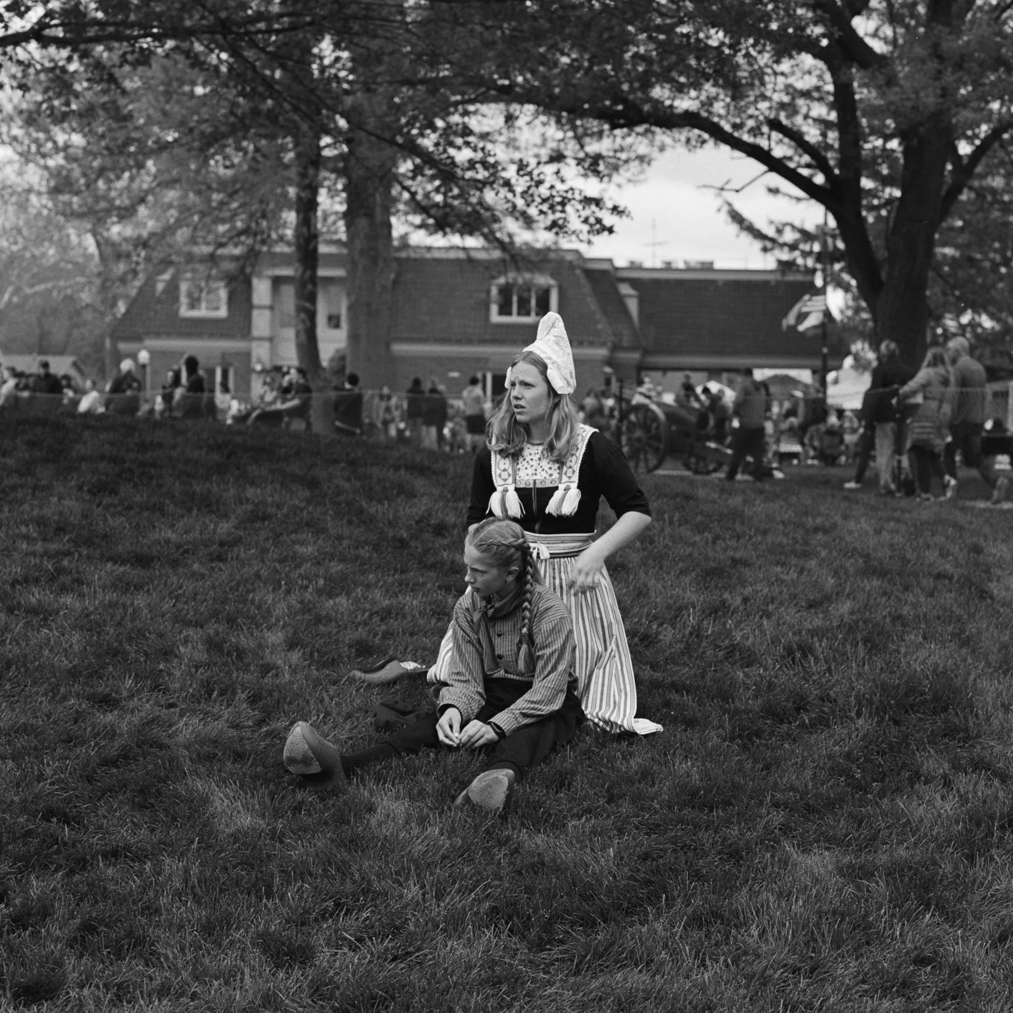 A woman dressed in traditional clothing, standing beside a girl sitting on the grass, in a park with a crowd of people and trees in the background. Pella, Iowa Tulip Time Festival, Dutch Heritage