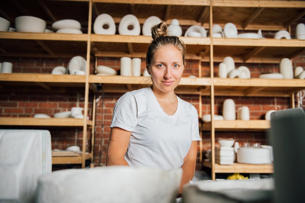 Woman pottery artist in white t-shirt smiling in pottery studio with shelves of unglazed pottery.