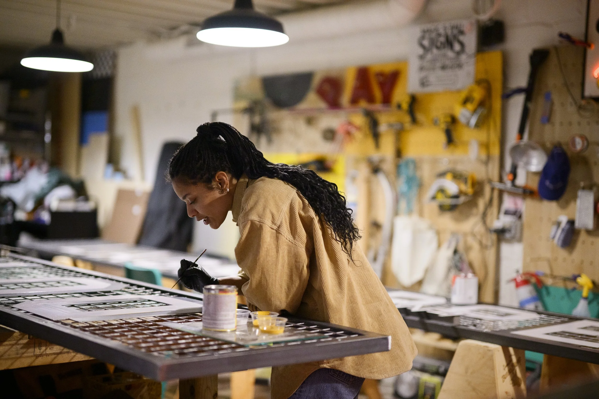 An artist painter sign designer woman with long curly hair wearing a beige jacket is working on art projects in a workshop, surrounded by tools and supplies on a workbench and wall.