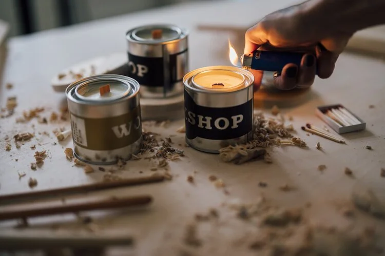 A person lighting a candle in a can with a "WASHOP" label, surrounded by other candles and scattered wax and debris on a table.