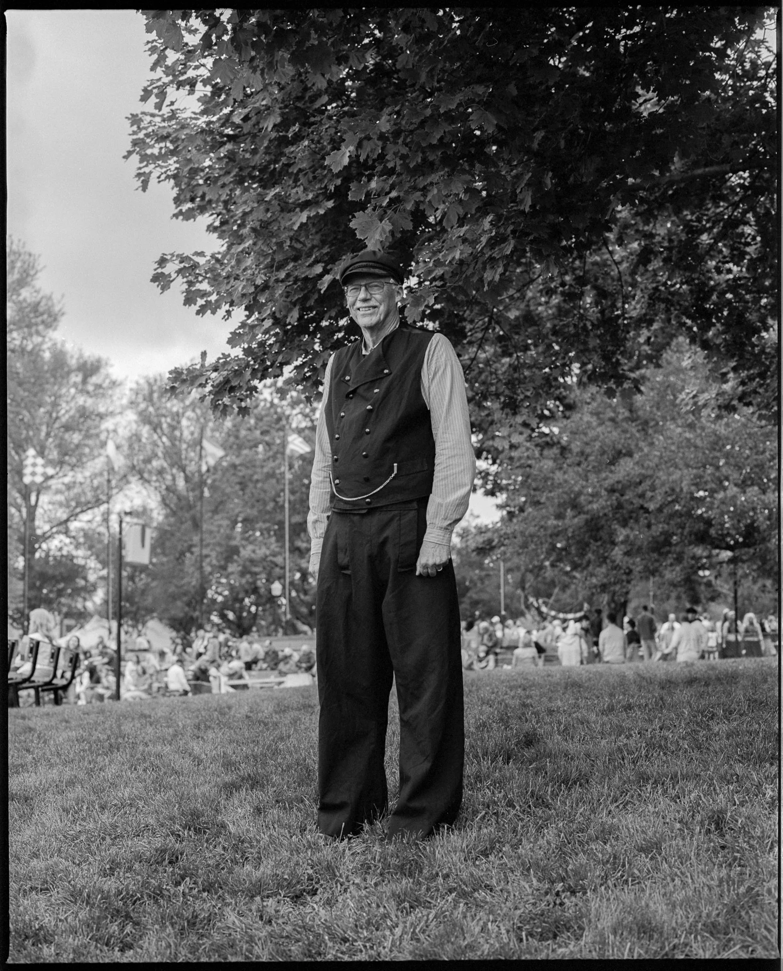A smiling elderly man wearing a cap, glasses, a double-breasted vest, and loose pants standing on grass in a park with trees and people in the background. Pella, Iowa Tulip Time Festival, Dutch Heritage