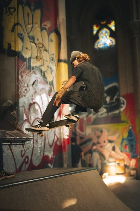 Skateboarder performing an aerial trick inside an indoor skate park with graffiti-covered walls and stained glass windows. Sk8 Liborius