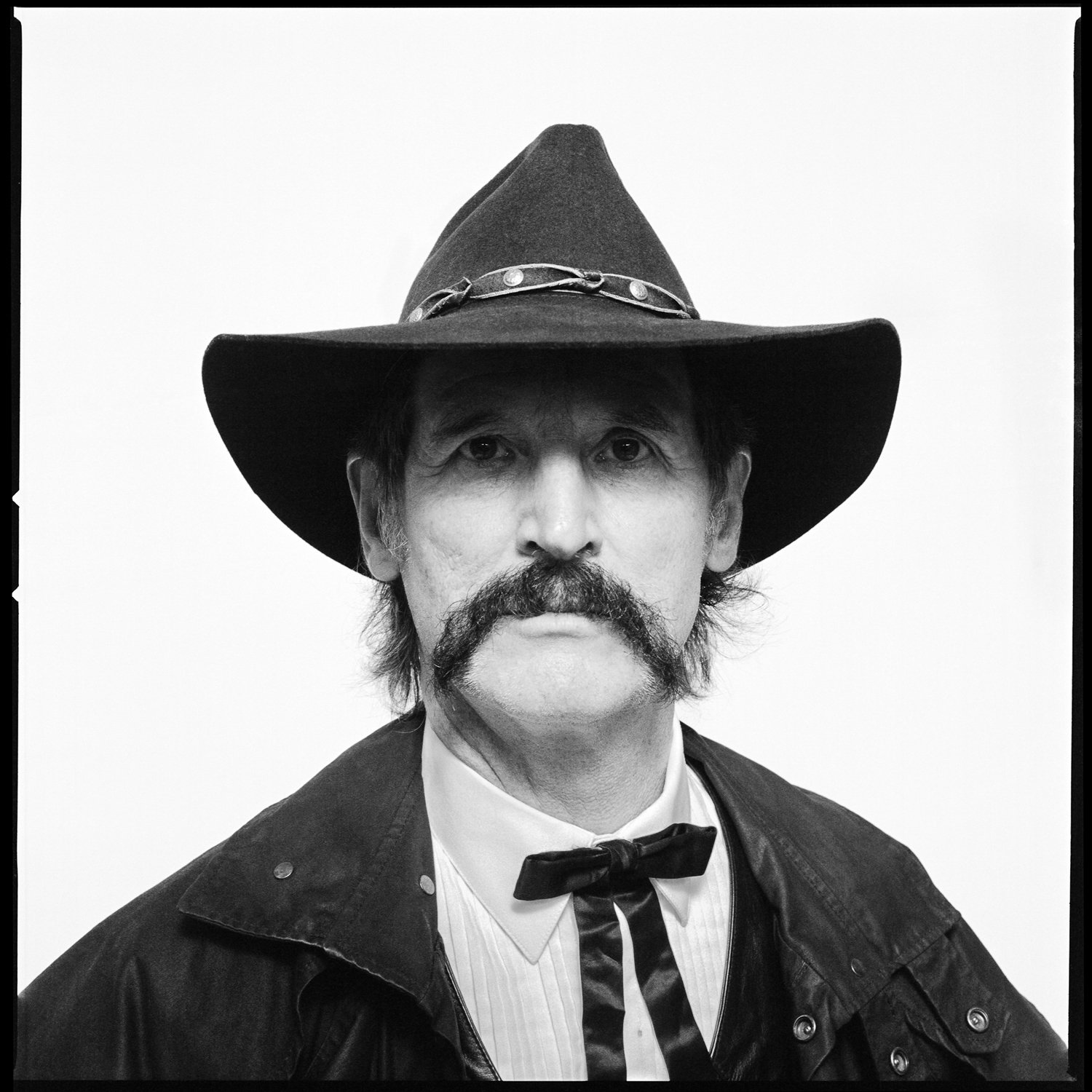 Black and white portrait of a man wearing a wide-brimmed hat with a decorative band, a leather jacket, a white shirt, and a bow tie, with a mustache and serious expression. Pella, Iowa Tulip Time Festival, Dutch Heritage, Wyatt Earp