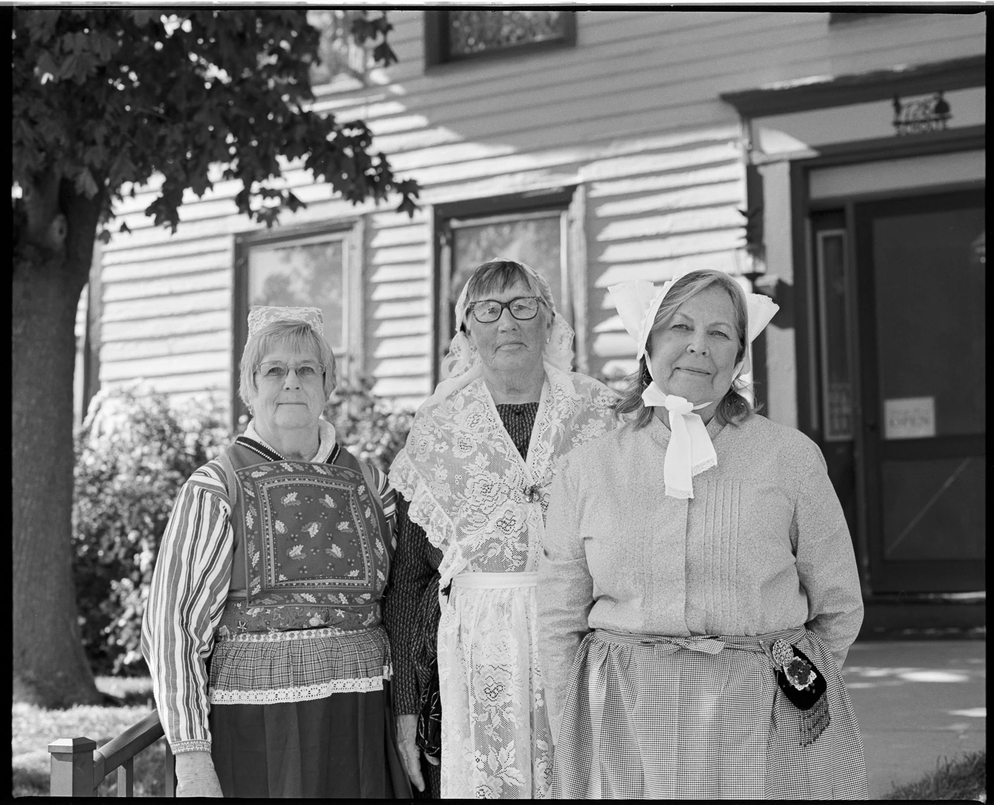 Three older women dressed in vintage clothing standing outdoors in front of a house, wearing accessories and traditional attire. Pella, Iowa Tulip Time Festival, Dutch Heritage
