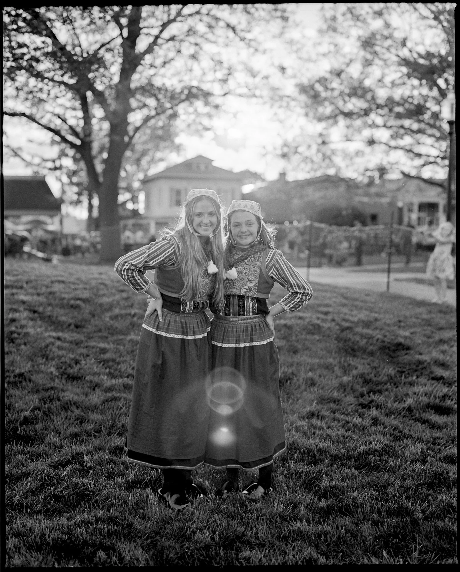 Two young girls wearing traditional outfits standing outdoors on grass, smiling, with sunlight filtering through trees in the background. Pella, Iowa Tulip Time Festival, Dutch Heritage