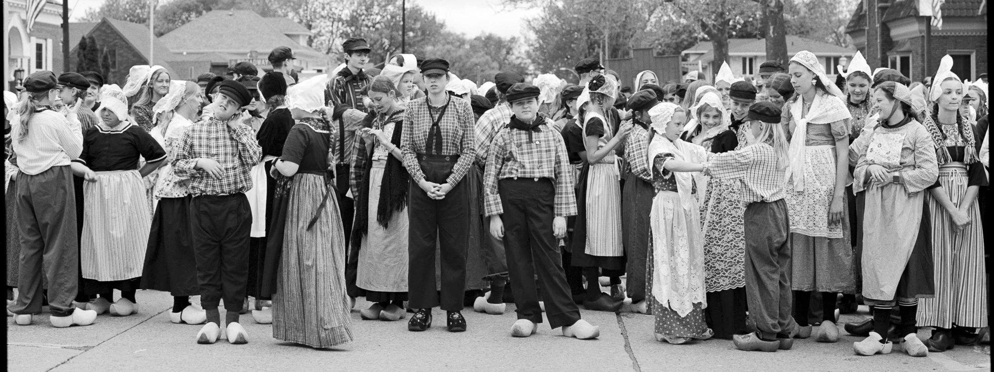 A large group of children dressed in historical clothing, possibly participating in a parade or festival, standing on a street with houses in the background. Pella, Iowa Tulip Time Festival, Dutch Heritage