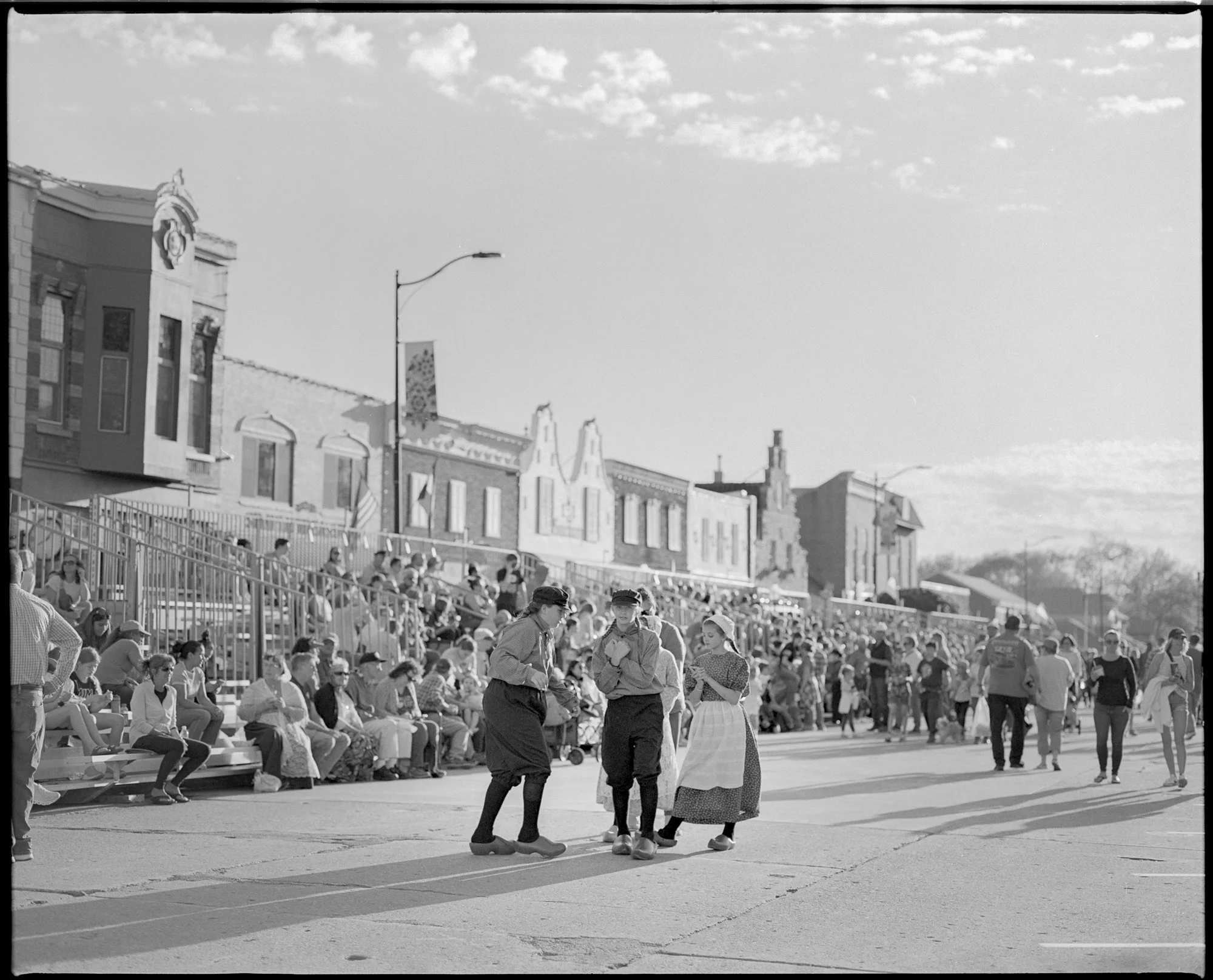A black and white photo of a crowd gathering on a street with historic-style buildings in the background. People dressed in vintage clothing suggesting a historical or cultural event. Pella, Iowa Tulip Time Festival, Dutch Heritage