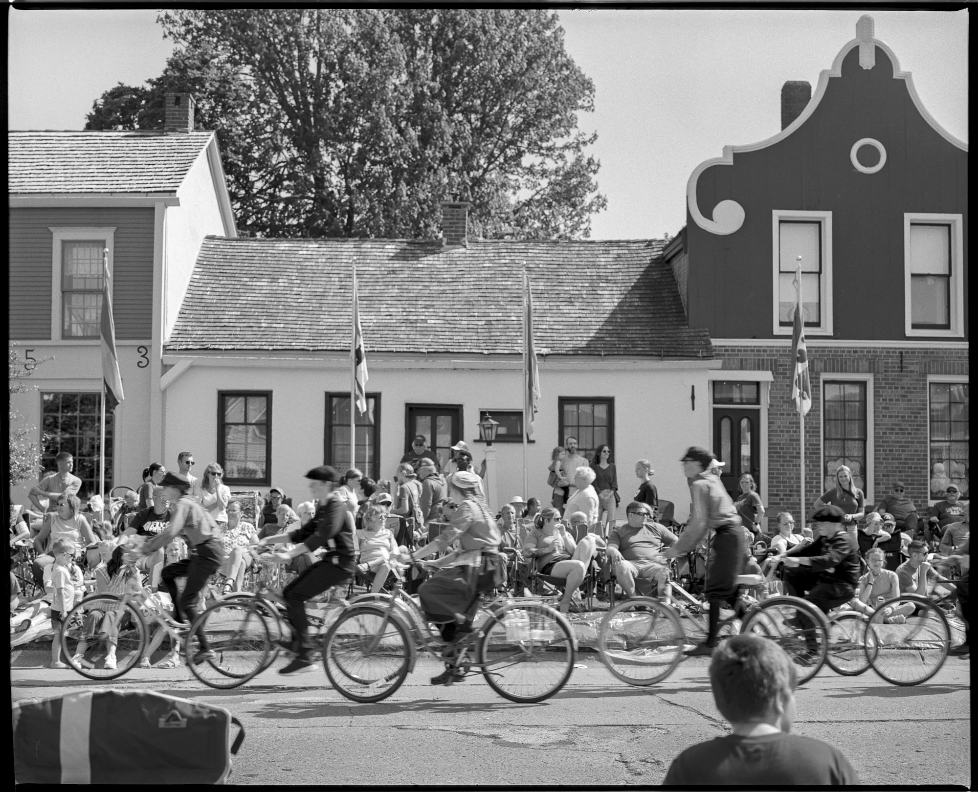 A black and white photo of a parade with people riding bicycles on a street and an audience watching from the sidewalk, in front of buildings with flags, trees, and a clear sky. Pella, Iowa Tulip Time Festival, Dutch Heritage