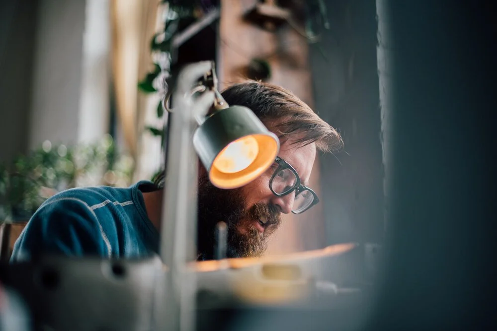 A man with glasses and a beard working at a desk, illuminated by a desk lamp, with a focused expression.