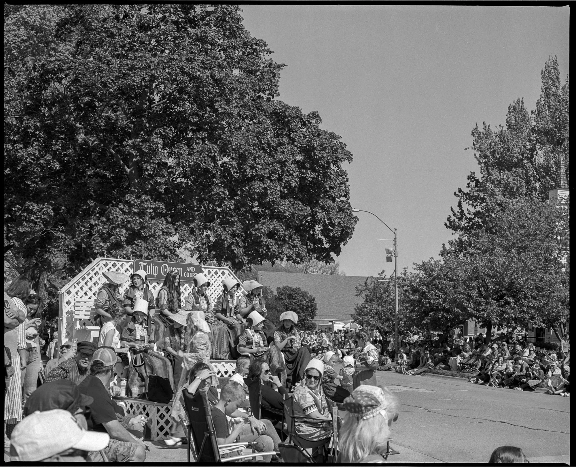 Black and white photo of a crowd gathered for a parade, with people sitting and standing along the street, and a float labeled 'Tulip Queen and Court' with women in costumes and large hats. Pella, Iowa Tulip Time Festival, Dutch Heritage