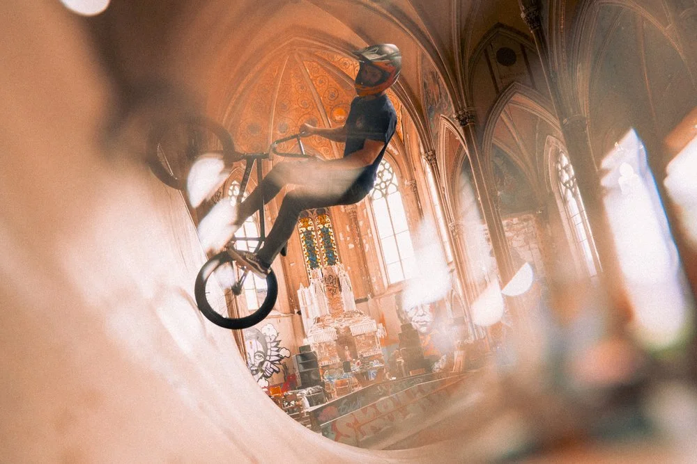 A person riding a BMX bike inside a church with arched windows and stained glass, wearing a helmet and black clothing. Sk8 Liborius