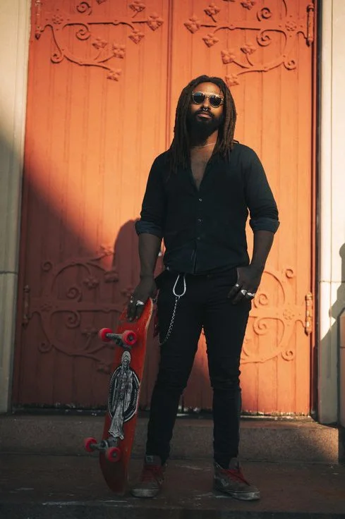 A man with dreadlocks and sunglasses standing in front of an orange metal door holding a skateboard. Sk8 Liborius
