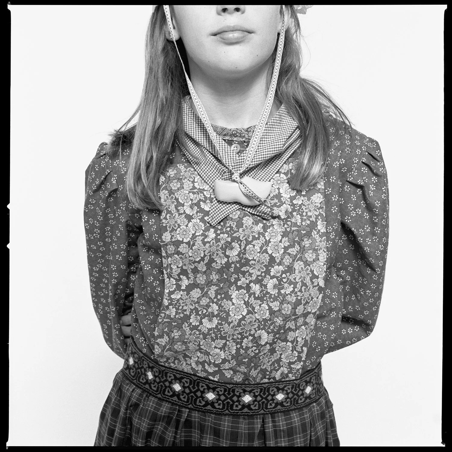 A young girl in a floral embroidered blouse and plaid skirt, wearing a bow tie and a patterned scarf, standing with her hands behind her back against a plain background. Pella, Iowa Tulip Time Festival, Dutch Heritage