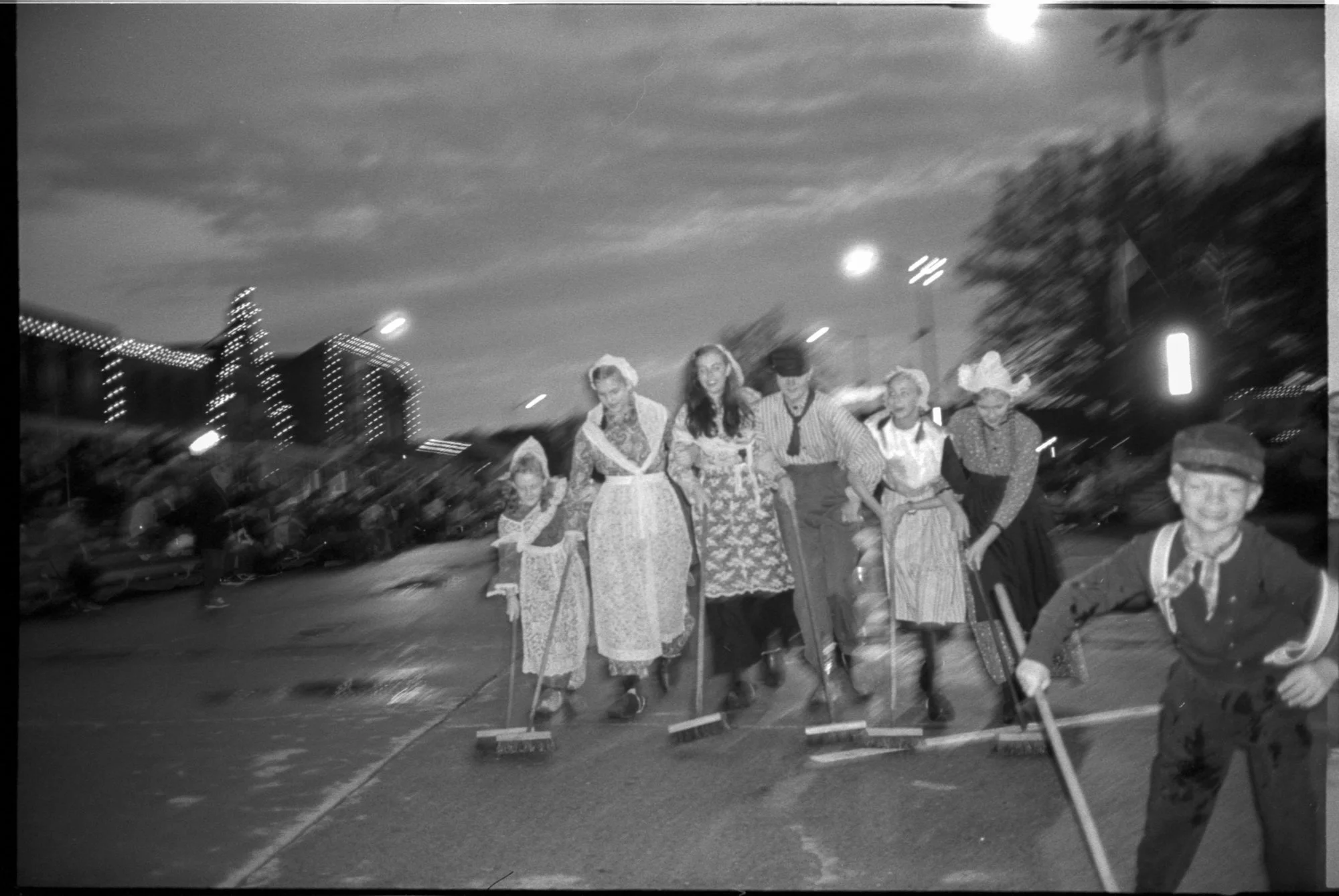 Group of people dressed in traditional or vintage costumes, smiling and walking on a street at night, with some holding brooms or sticks. Pella, Iowa Tulip Time Festival, Dutch Heritage