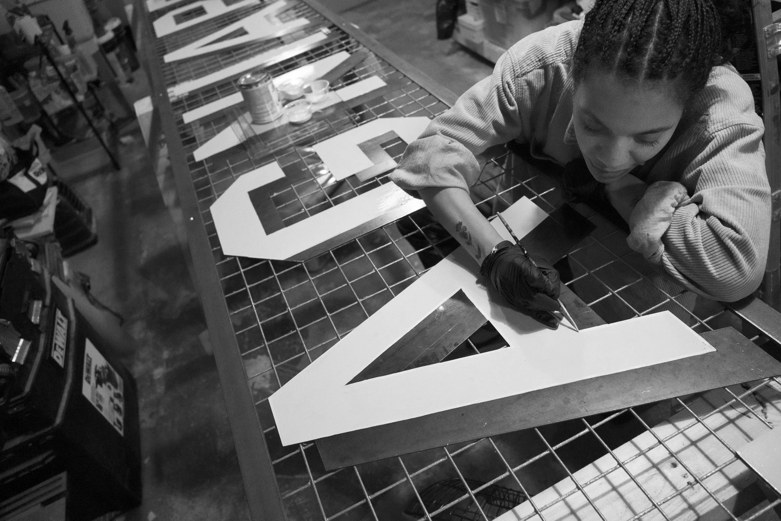 An artist sign maker painter is working on a large sign, painting or drawing on a sheet of paper at a workshop or art studio. The sign appears to be a letter or number with a black outline, lying on a table covered with a grid pattern.
