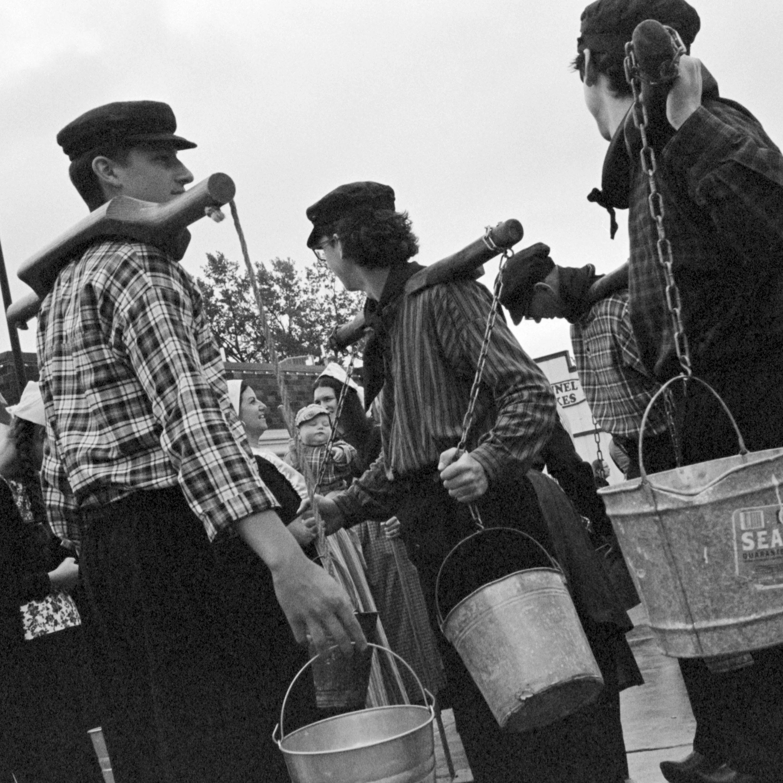Group of children wearing plaid shirts and berets at an outdoor event, holding buckets, with some children smiling and talking. Pella, Iowa Tulip Time Festival, Dutch Heritage
