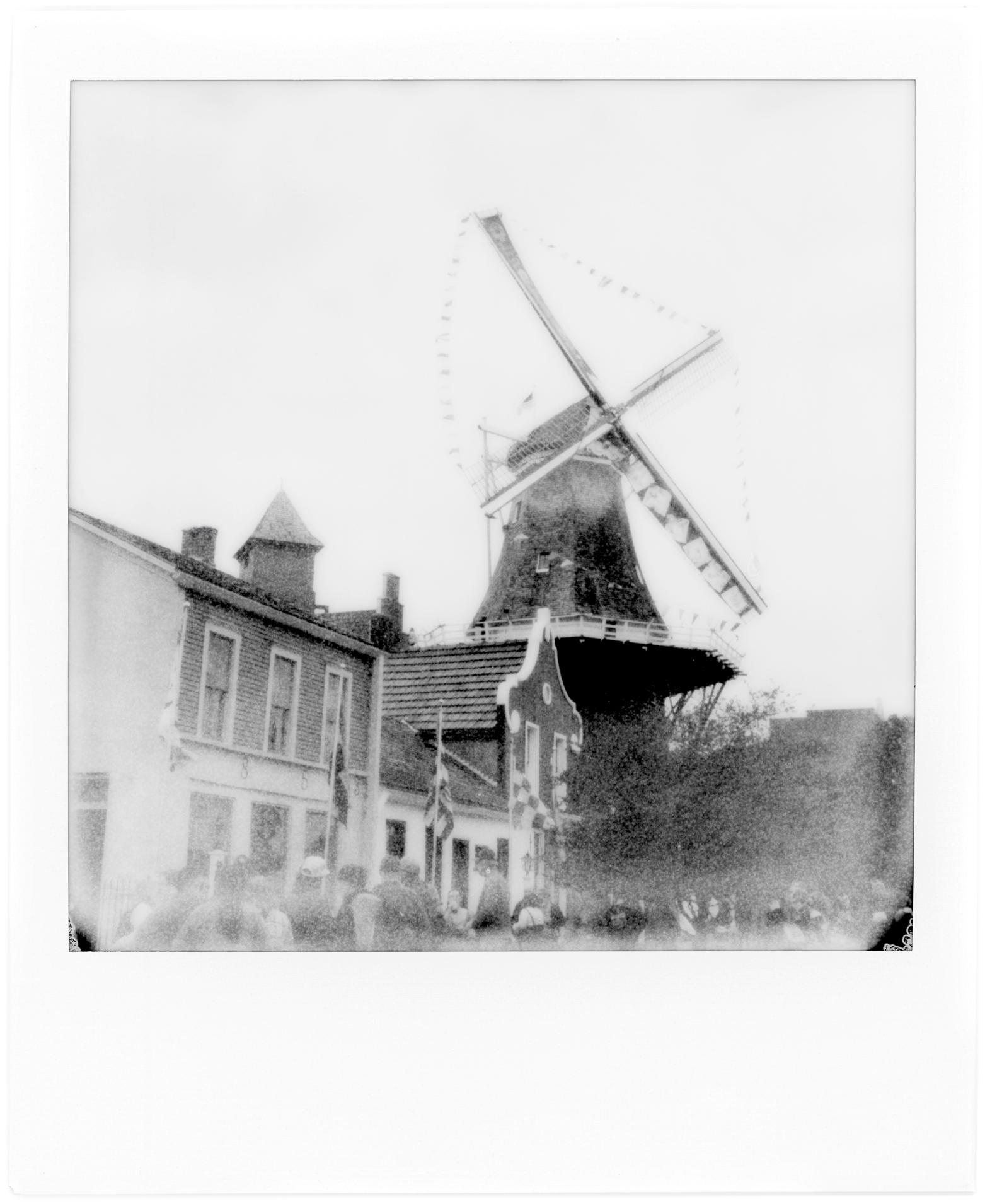 Black and white photo of a traditional Dutch windmill surrounded by historic buildings, with a crowd of people in the foreground. Pella, Iowa Tulip Time Festival, Dutch Heritage
