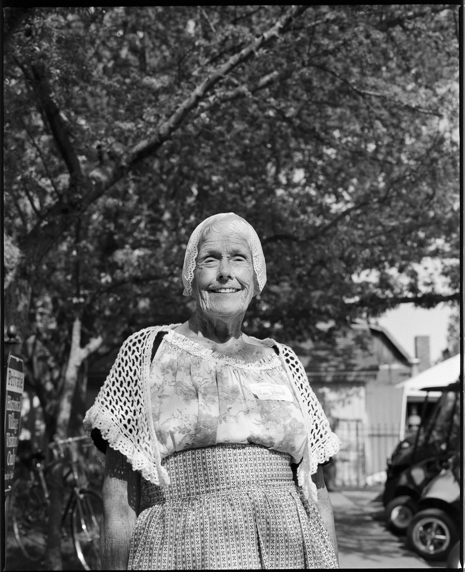 A smiling elderly woman wearing vintage clothing standing outdoors with trees and parked vehicles in the background. Pella, Iowa Tulip Time Festival, Dutch Heritage