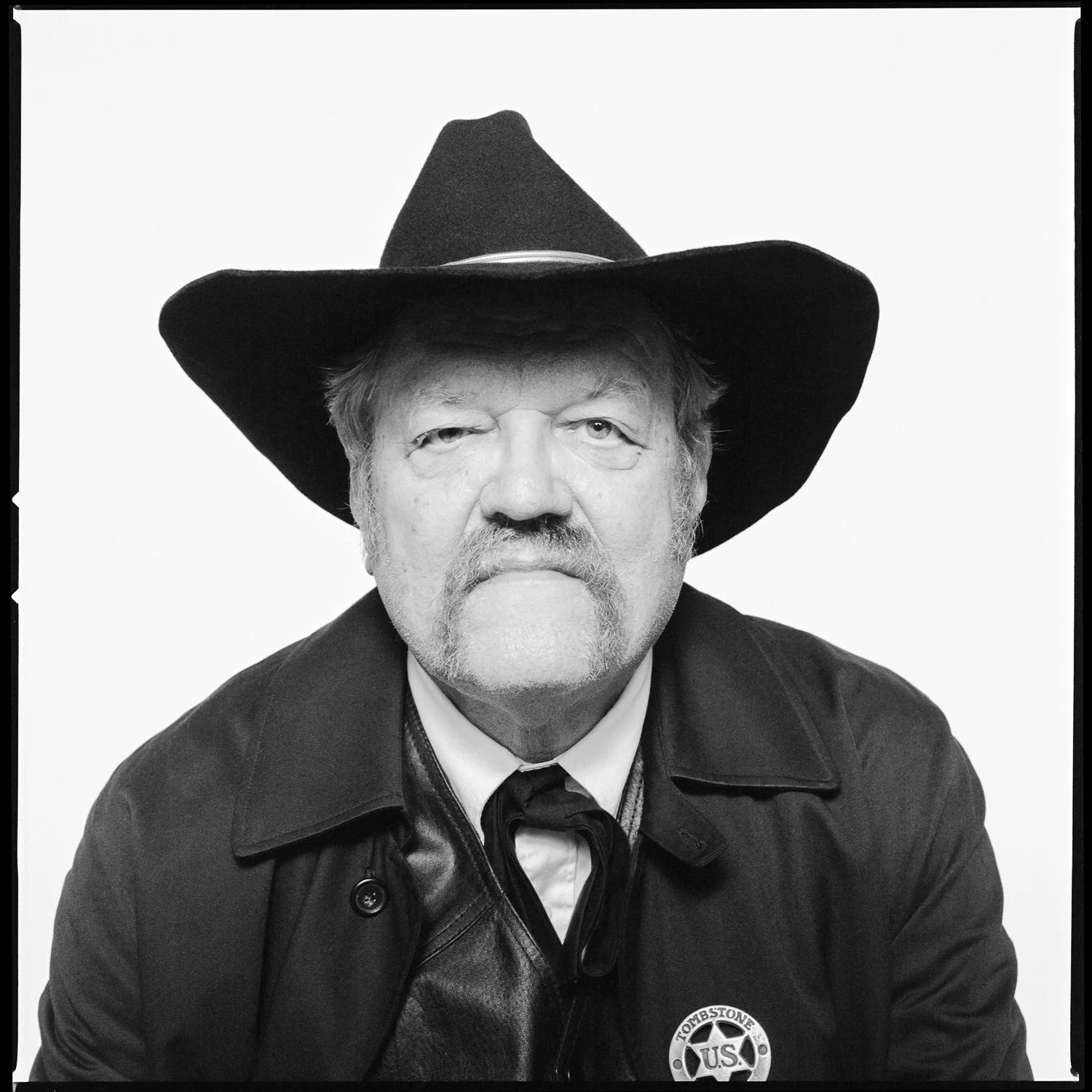 Black and white portrait of an older man wearing a cowboy hat, leather jacket, and a badge that says 'Tomstone U.S.' with a serious expression. Pella, Iowa Tulip Time Festival, Dutch Heritage, Wyatt Earp