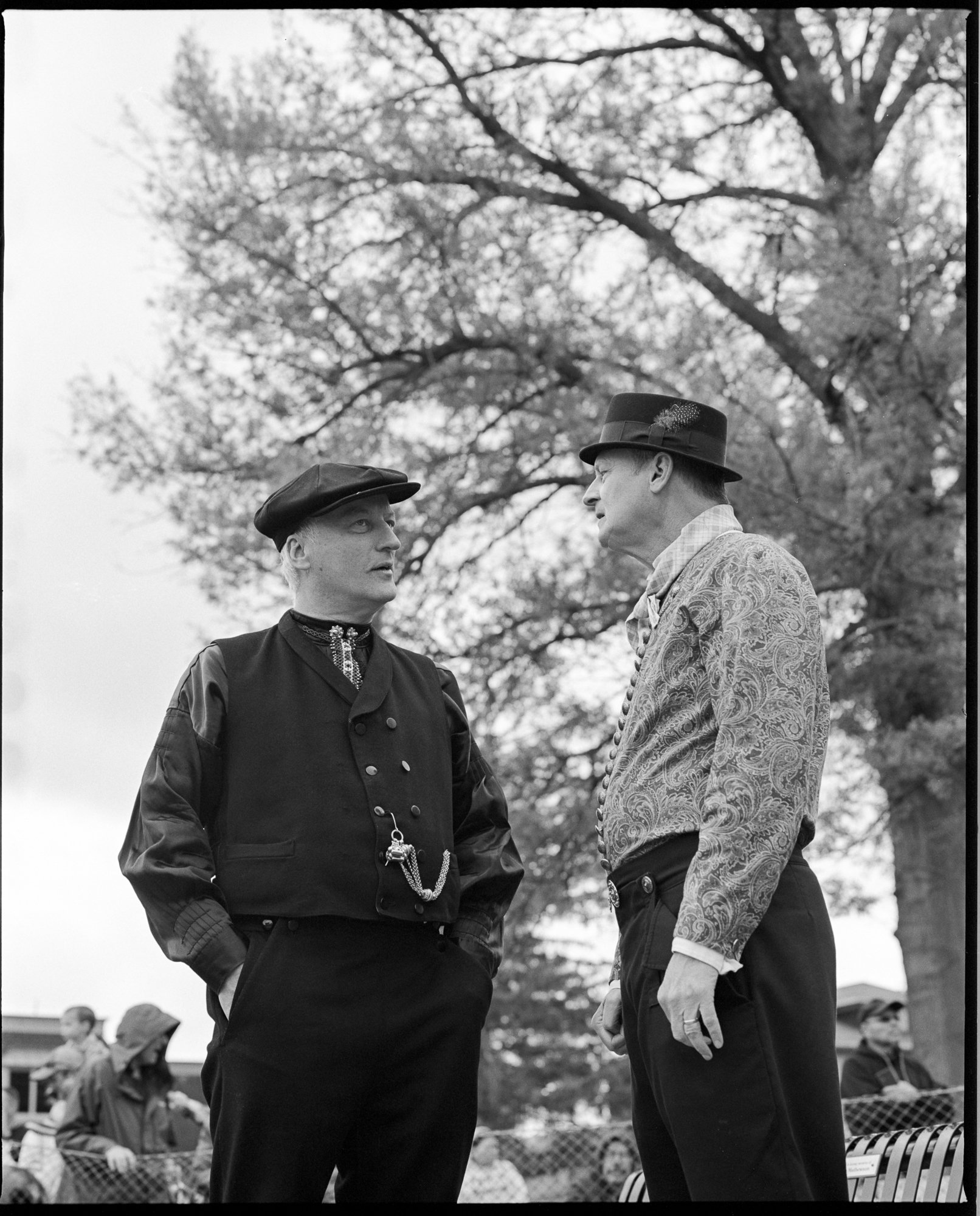Two men dressed in vintage clothing are standing and talking outdoors in front of a large tree, with a crowd of people in the background. Pella, Iowa Tulip Time Festival, Dutch Heritage