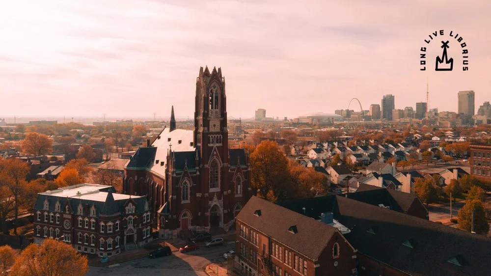 Aerial view of a cityscape during fall, featuring a large historic church with a tall steeple in the foreground and a mix of residential and modern buildings in the background, with the St. Louis Arch visible on the horizon. Sk8 Liborius
