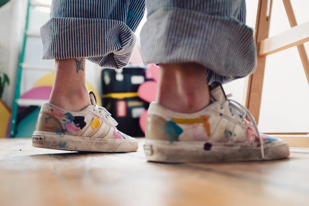 Close-up of a artist's painted sneakers and rolled-up jeans, standing on a wooden floor indoors.