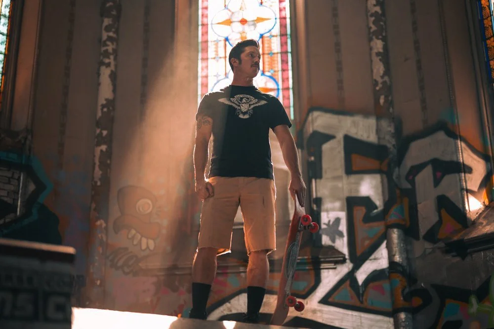A man standing with a skateboard inside a graffiti-covered skate shop or lounge, with colorful murals and a stained glass window in the background. Sk8 Liborius