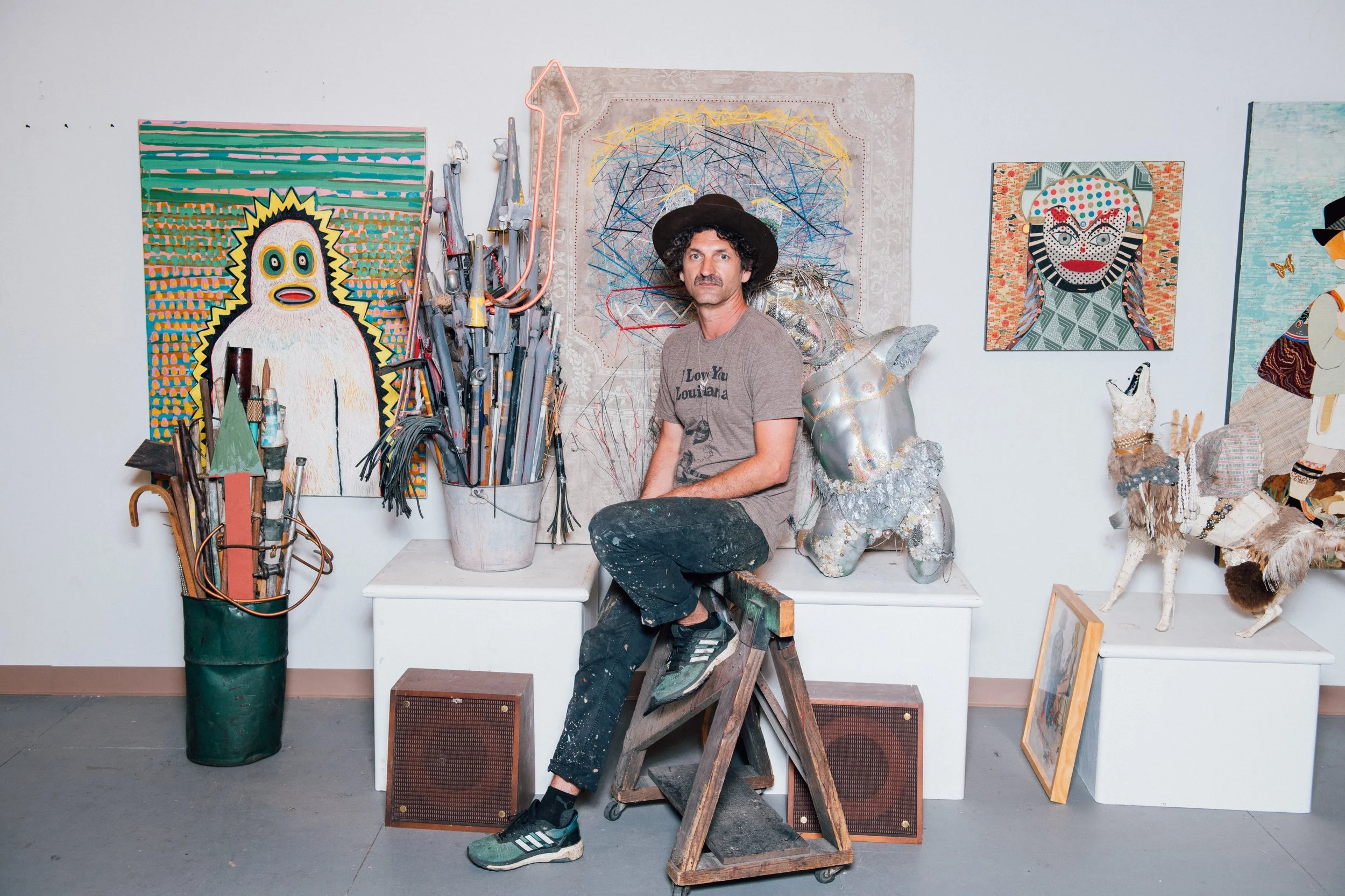 An artist's studio with a male artist sitting on a wooden stool surrounded by abstract and colorful paintings, sculptures, and art supplies.