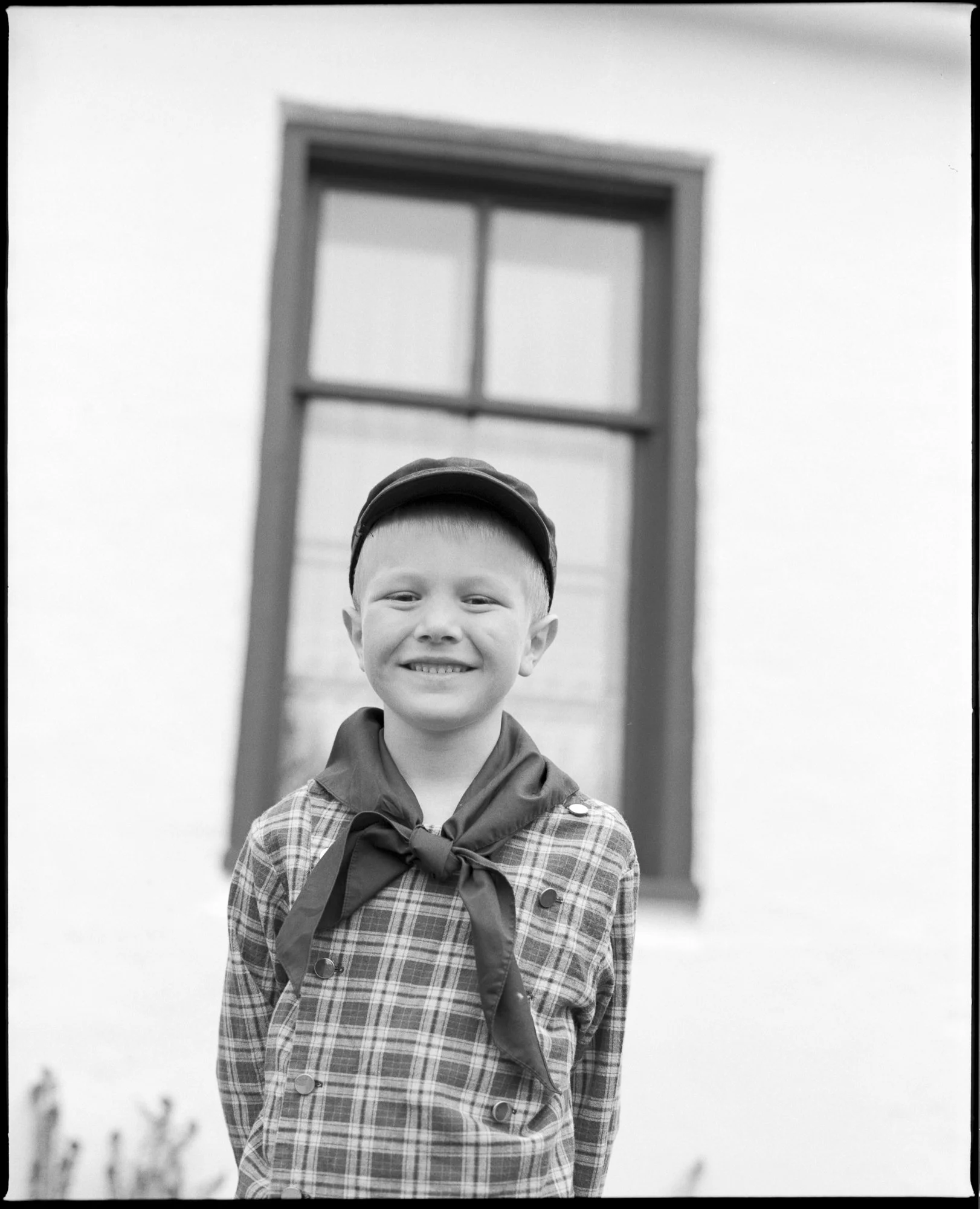 A young boy in a plaid shirt and neckerchief, smiling while standing outdoors in front of a white wall with a window. Pella, Iowa Tulip Time Festival, Dutch Heritage