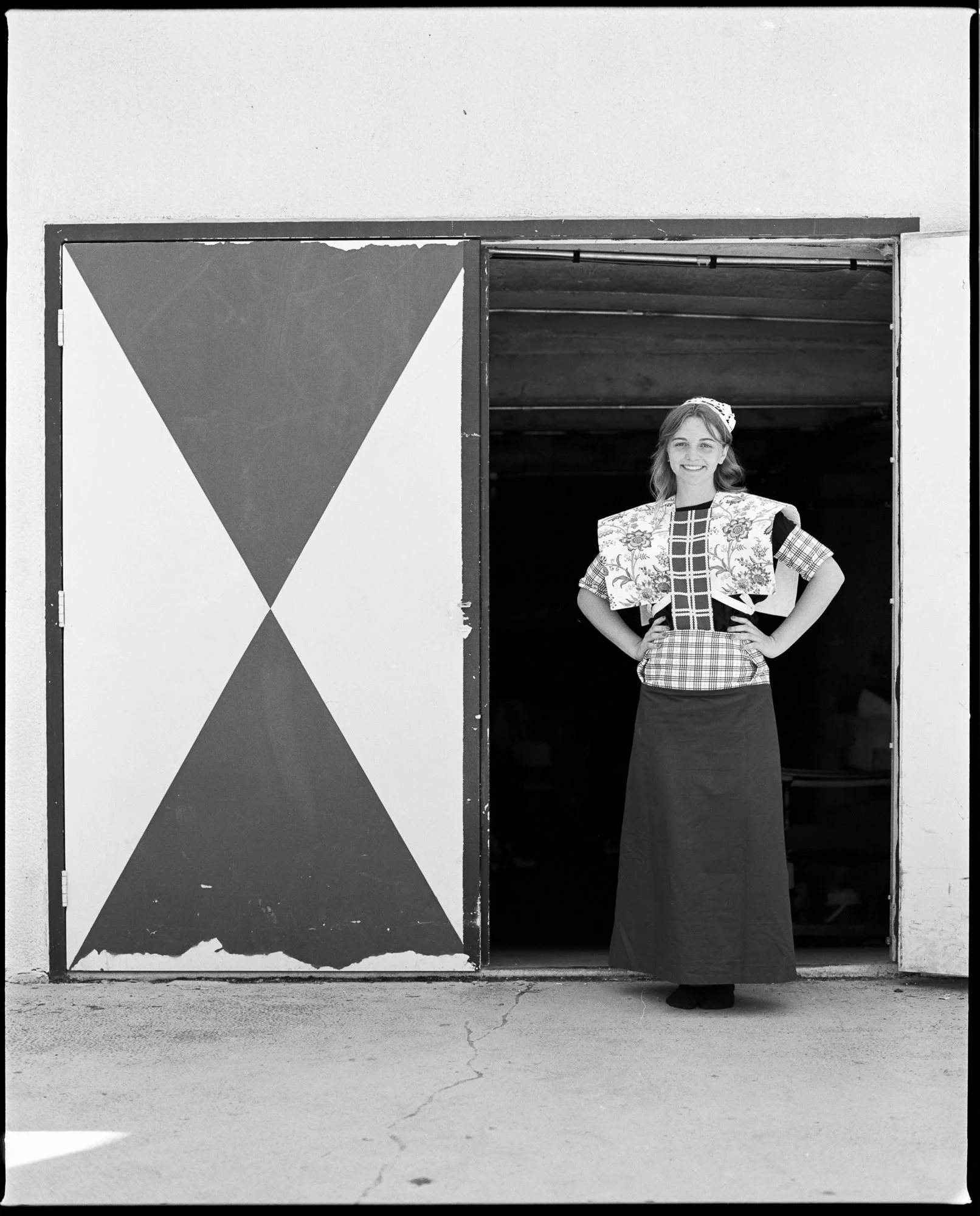 A woman stands in a doorway, smiling and wearing a patterned blouse, a long skirt, and a headscarf, with her hands on her hips, in front of a dark interior. Pella, Iowa Tulip Time Festival, Dutch Heritage