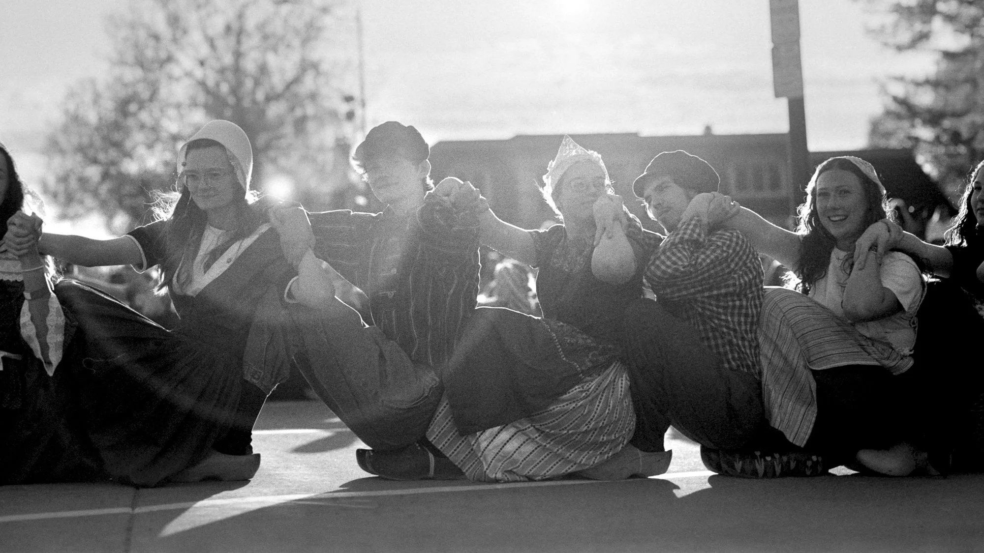 Group of children dressed in traditional costumes holding hands and sitting on the ground outdoors, backlit by sunlight. Pella, Iowa Tulip Time Festival, Dutch Heritage