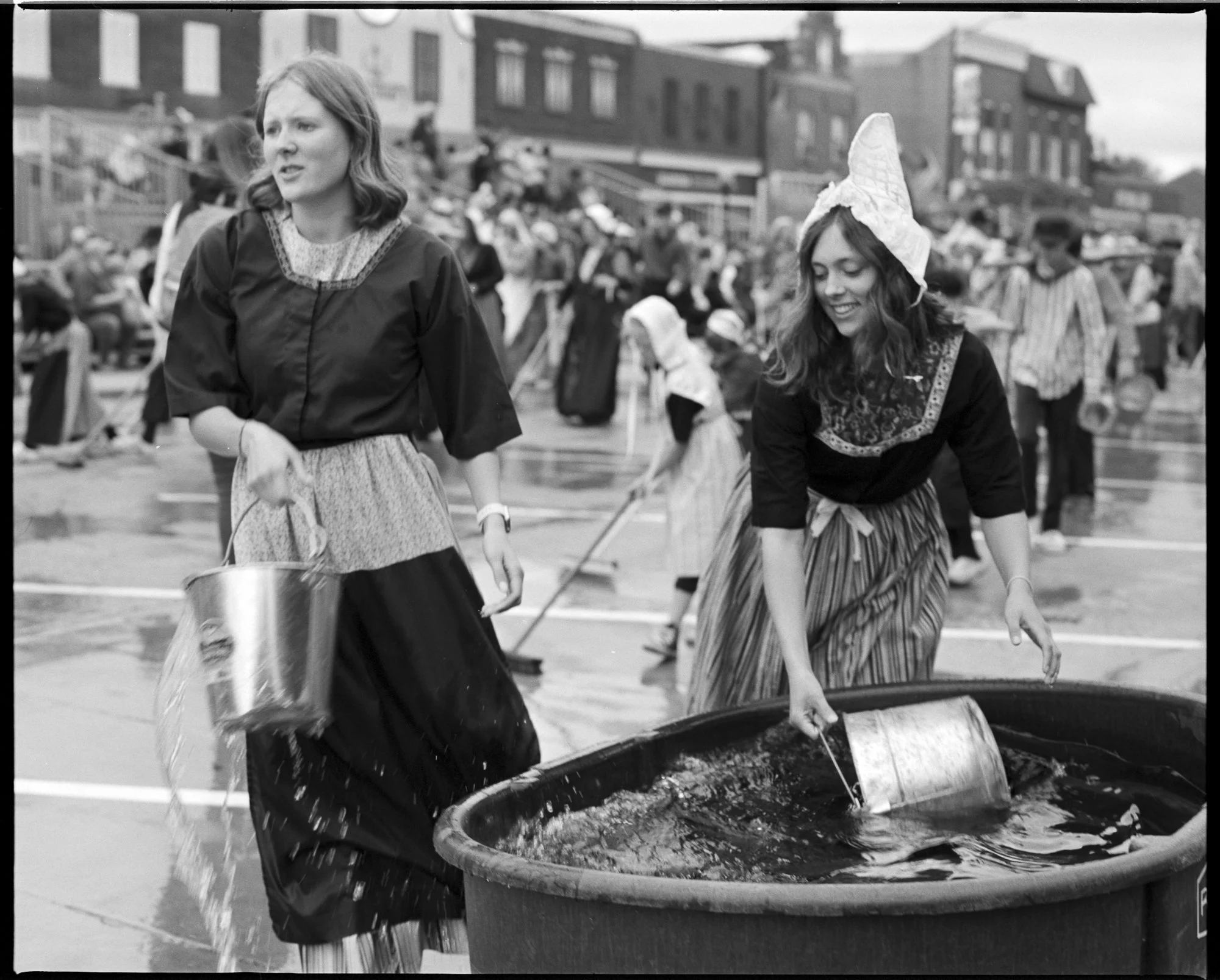 Two women dressed in old-fashioned clothing standing near large water barrel at an outdoor event, with people in the background. Pella, Iowa Tulip Time Festival, Dutch Heritage