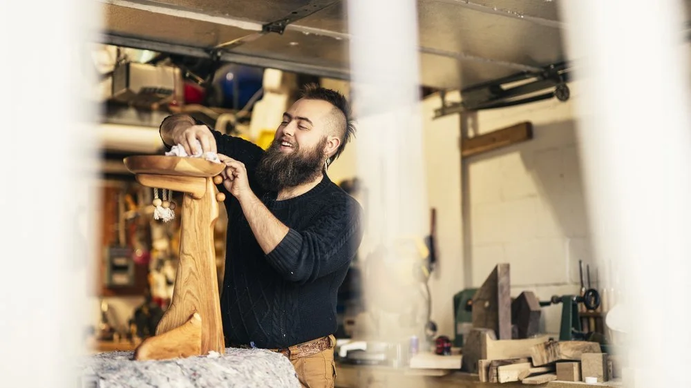 A bearded man furniture designer artist in a workshop, smiling, working on a piece of wood.