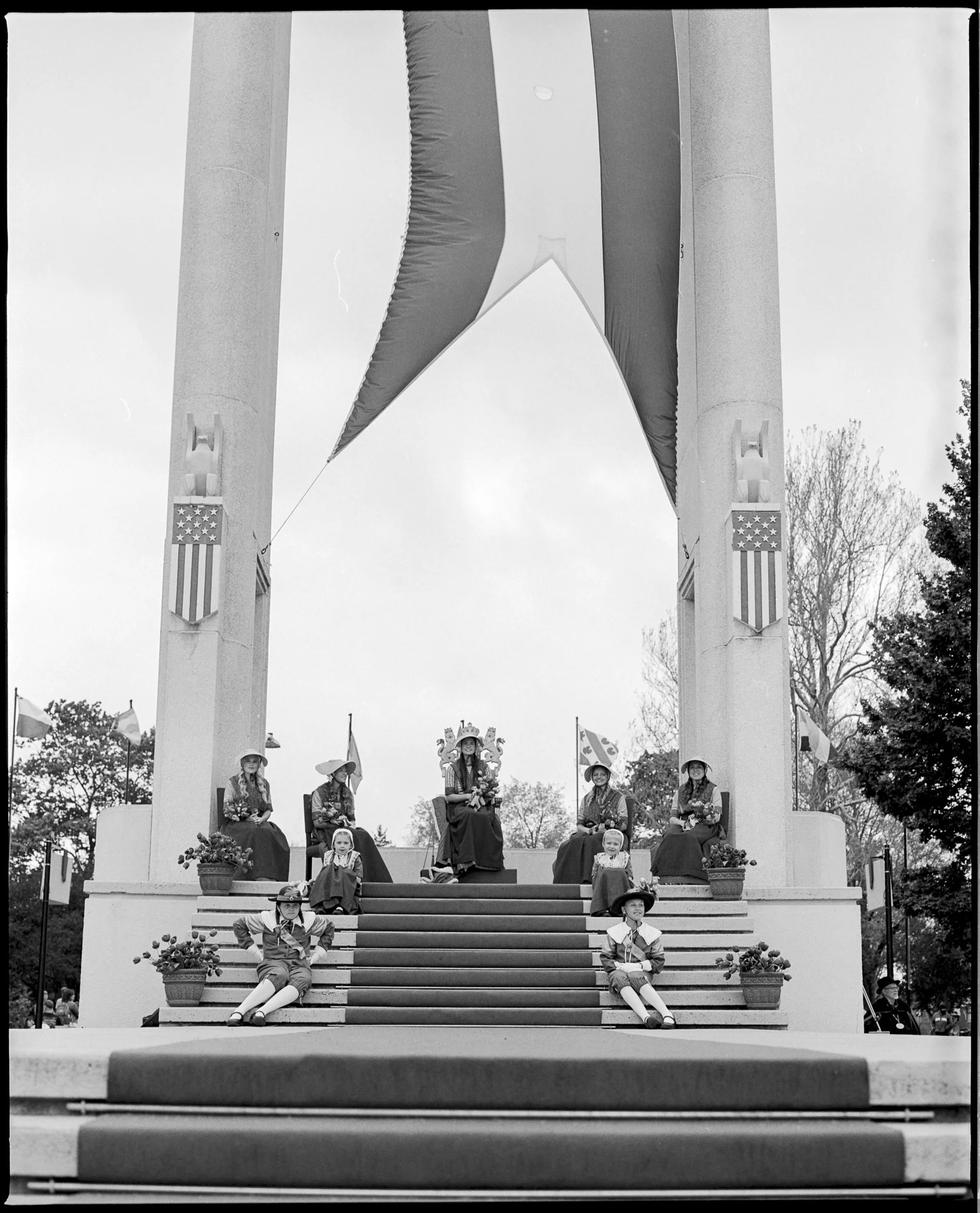 Black and white photo of a staged scene with people in vintage clothing sitting and standing on a decorated outdoor stage, with large flags hanging above and American flags on the columns. Decor includes potted plants on the steps. Pella, Iowa Tulip