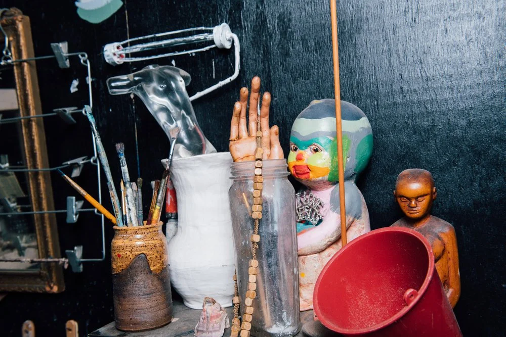 Assorted art and sculpture supplies including paintbrushes, jars, sculpted figurines, and decorative objects on a dark background.